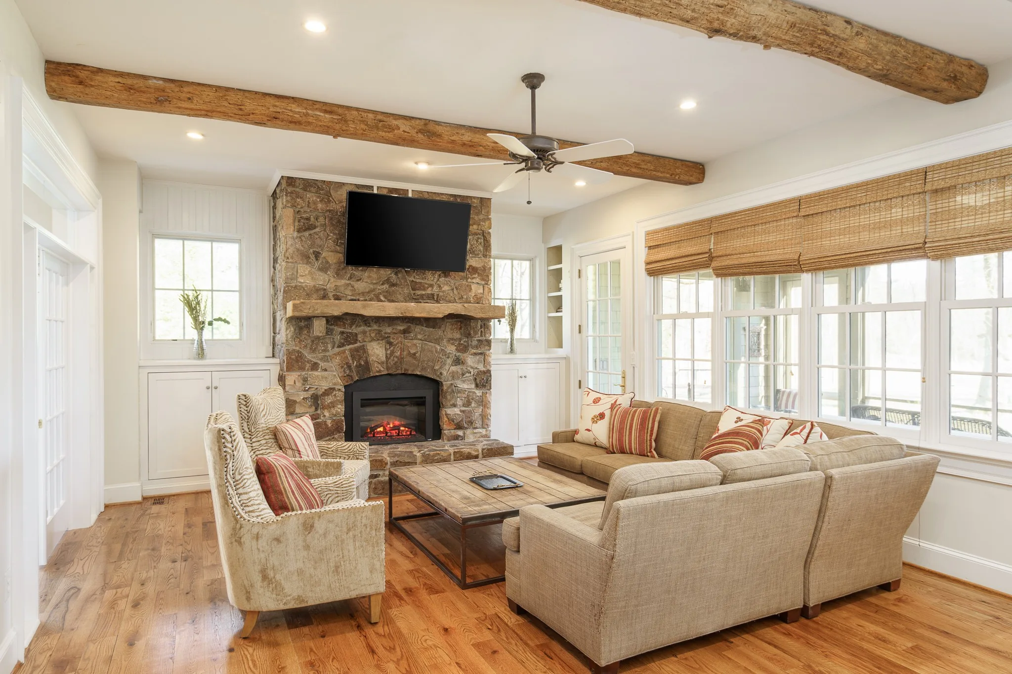 Living room with a stone fireplace, beige sofas, wooden ceiling beams and tall windows on two sides