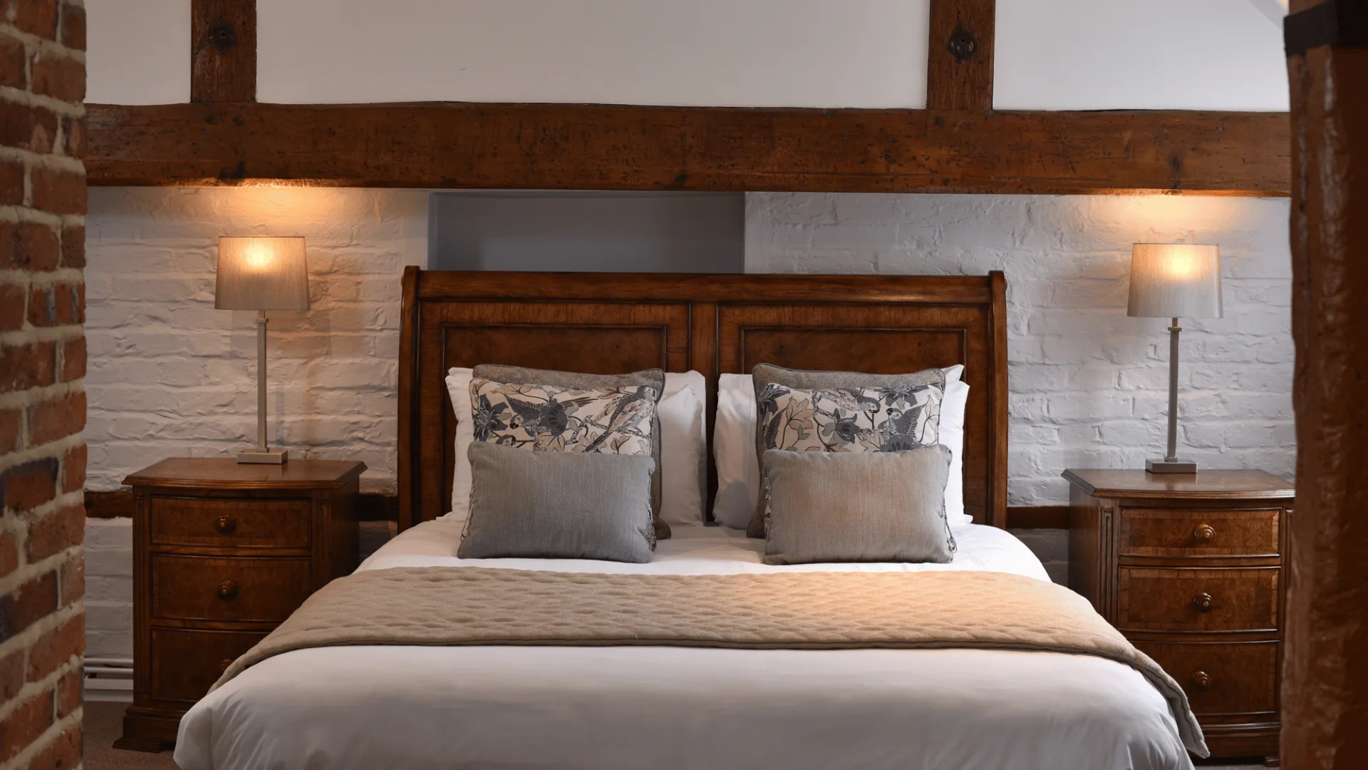 Double bed with patterned cushions between exposed timber beams, wooden bedside tables and wall lamps