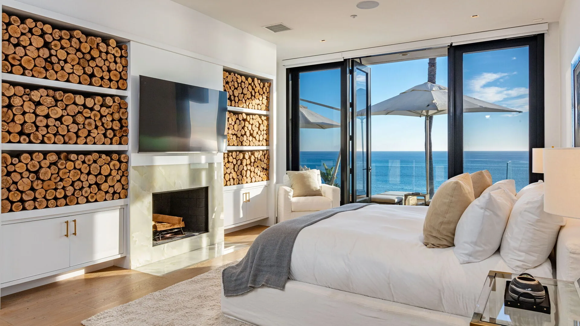 Bedroom with white bed, log stacked shelves around a fireplace and glass doors opening to the ocean