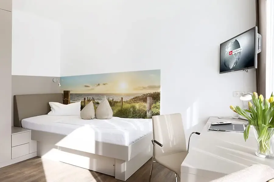 Bright single room with a white bed, a beach photo mural, a wall mounted TV and a desk with tulips