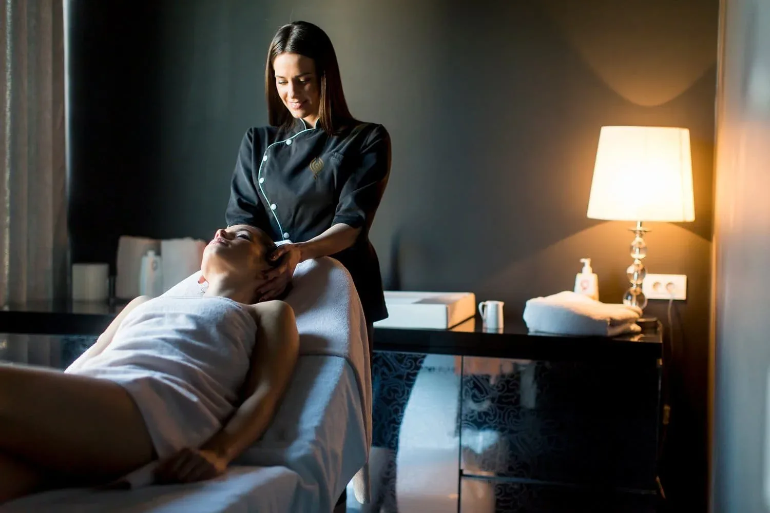 Dimly lit treatment room where a therapist gives a head massage to a client lying on a massage table