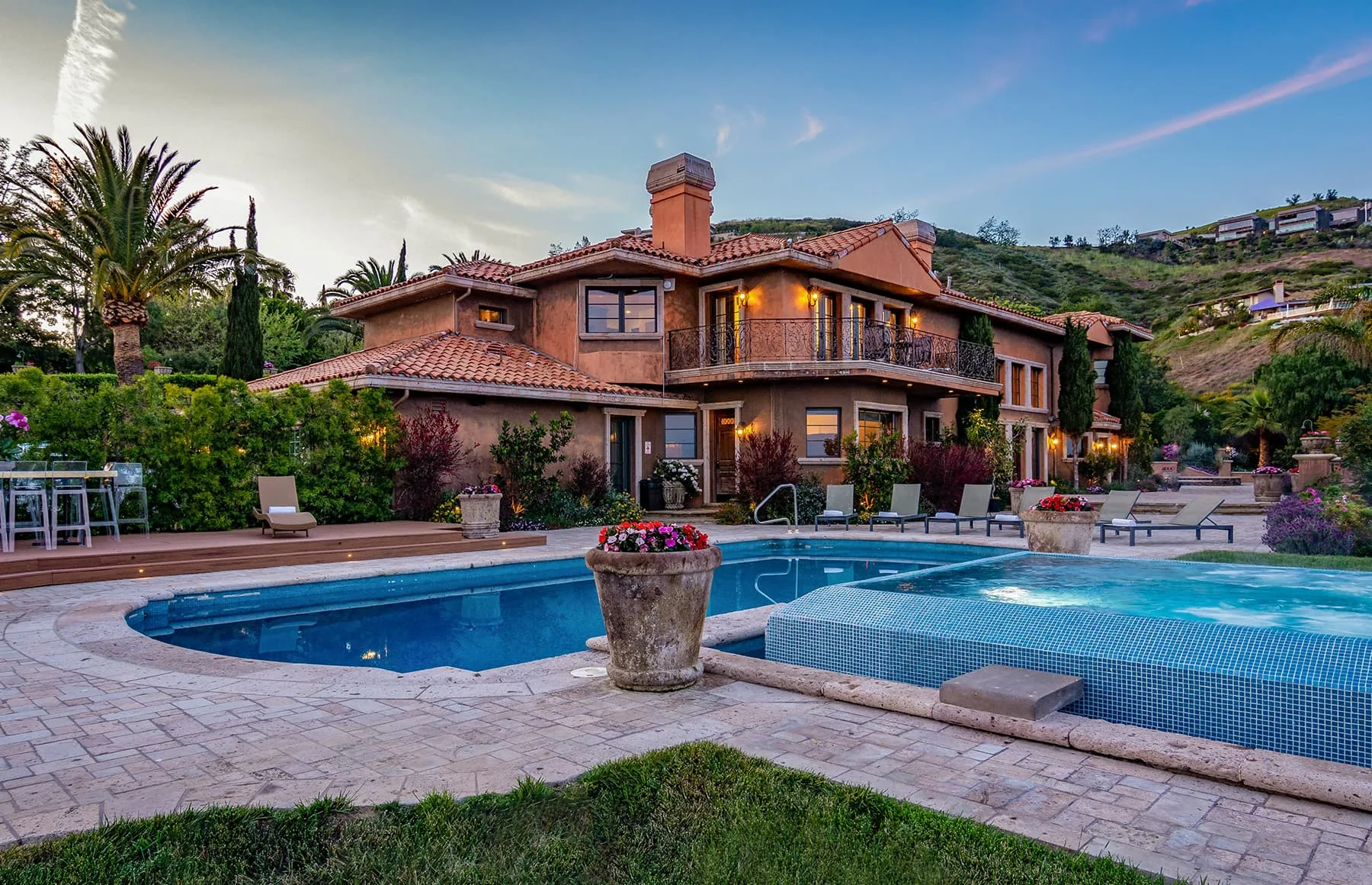 Mediterranean style house with a terracotta roof above a stone pool deck and hot tub at dusk