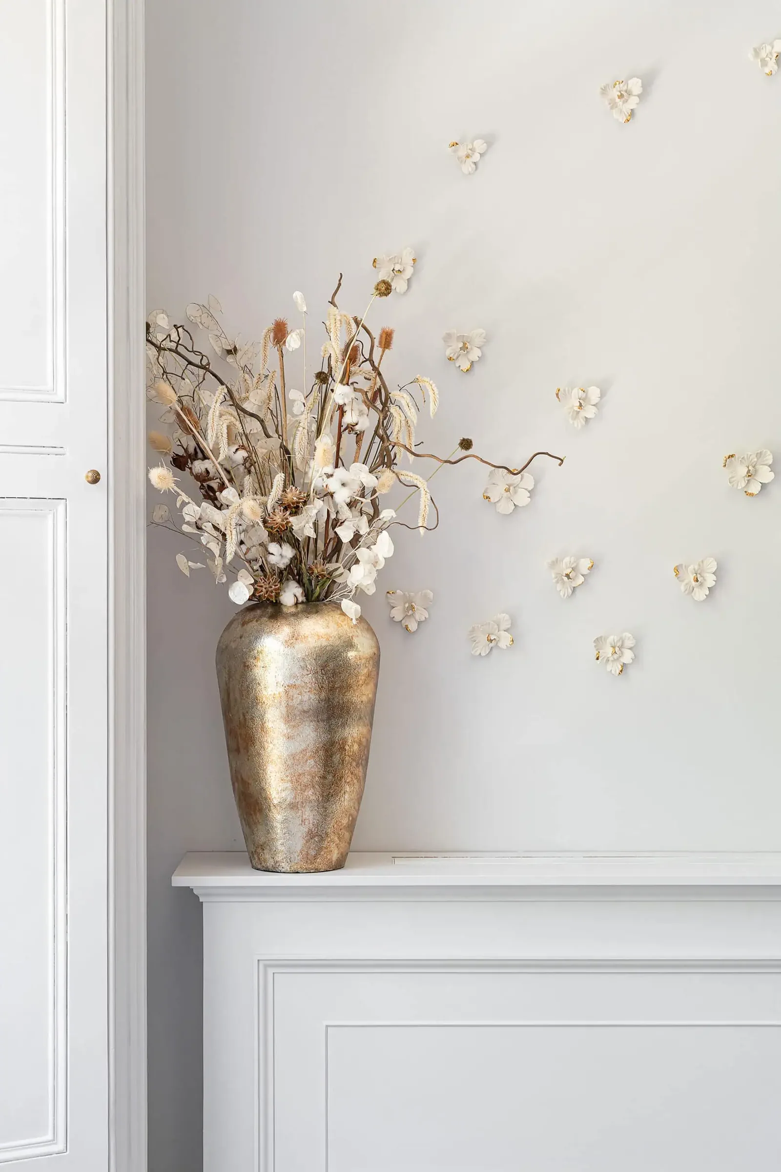 Gold vase of dried flowers on a white mantel below porcelain butterflies mounted on the wall