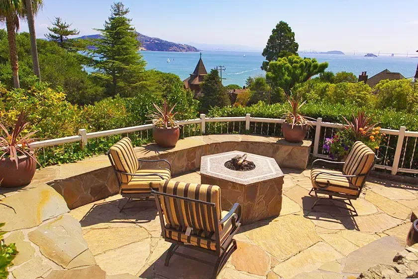 Flagstone garden patio with a fire pit and striped armchairs, trees and the bay beyond