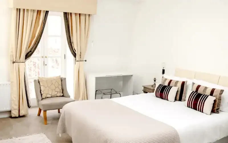 Bright bedroom with a double bed, striped cushions, an armchair and French windows with long curtains