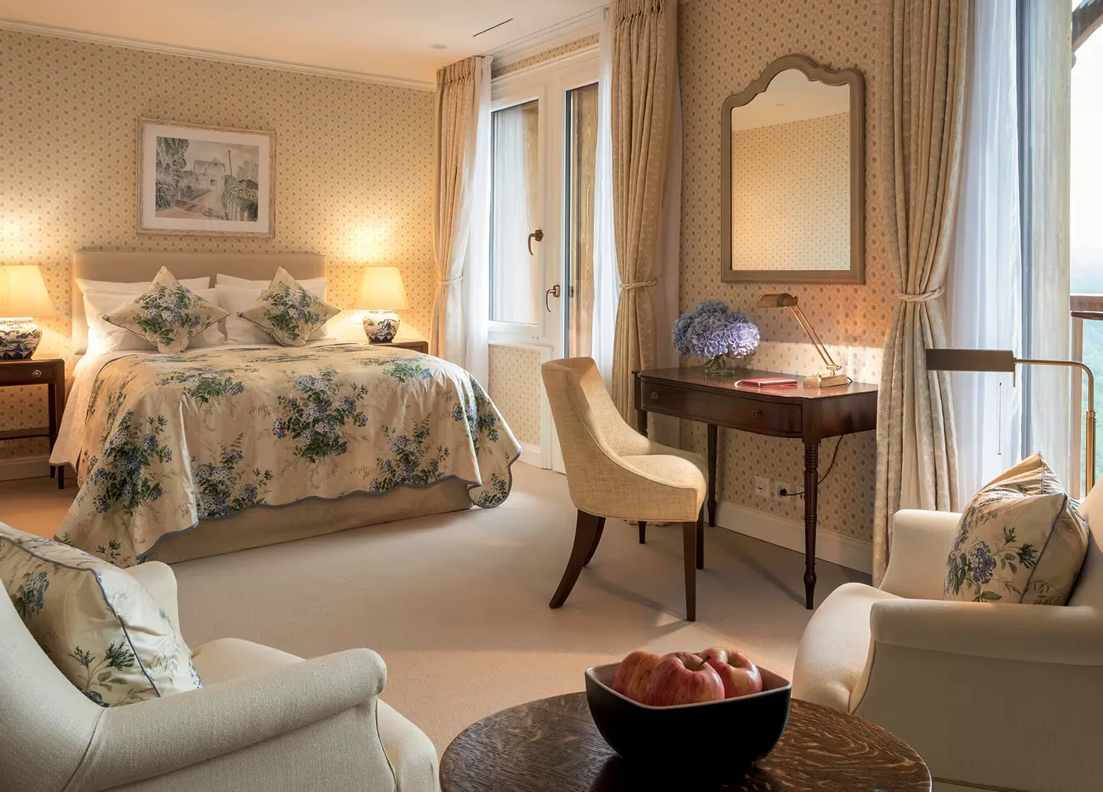 Bedroom with a floral covered bed, armchairs, a writing desk and tall curtained windows