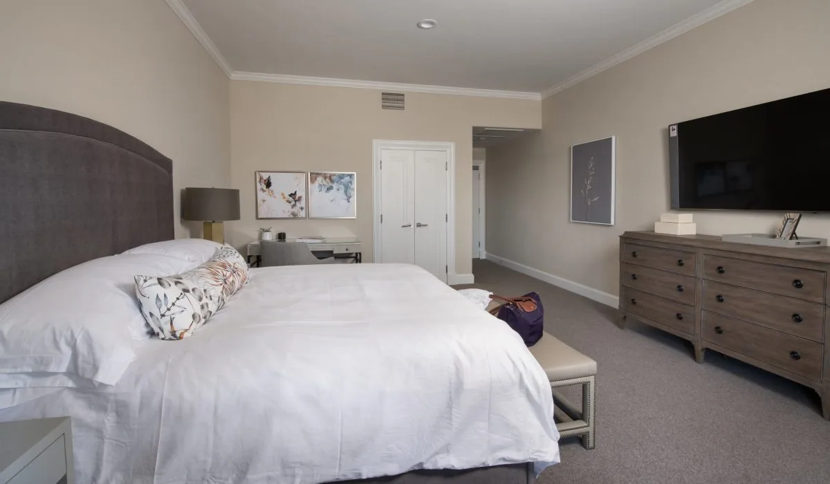 Private suite bedroom with a large white bed, upholstered headboard, dresser and wall television