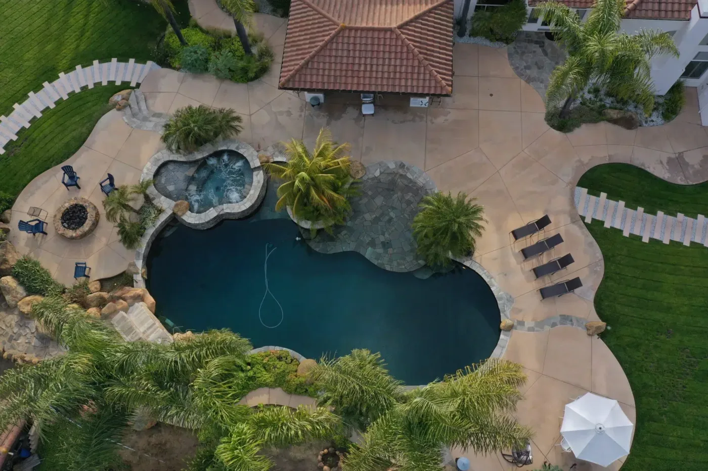Overhead view of a freeform pool and hot tub with sun loungers, palms and a lawn beside a house