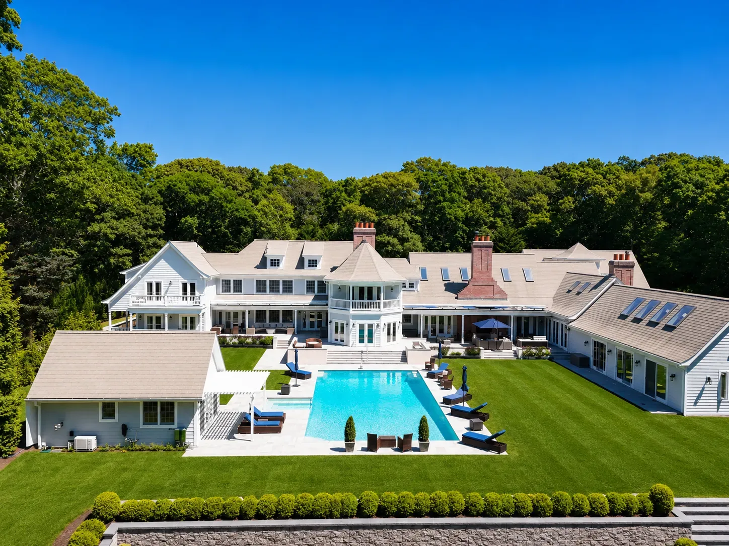 Aerial view of a shingle-style estate house with a rectangular pool, lawn and woodland behind
