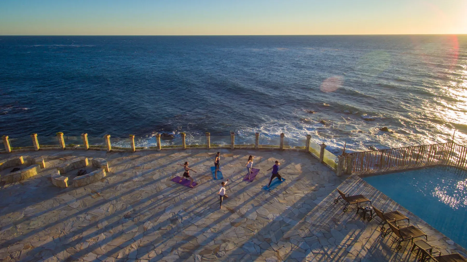Elevated view of a seaside terrace and pool at sunset with small figures practising yoga