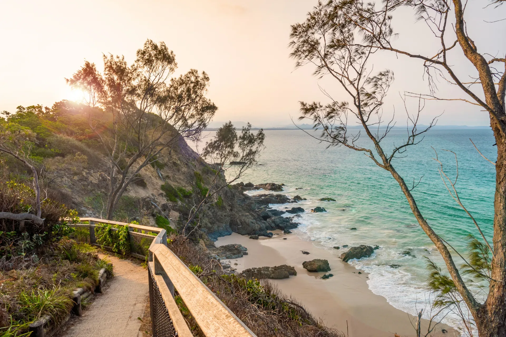 Timber walkway along a bush-covered headland above a sandy cove and turquoise sea at sunrise