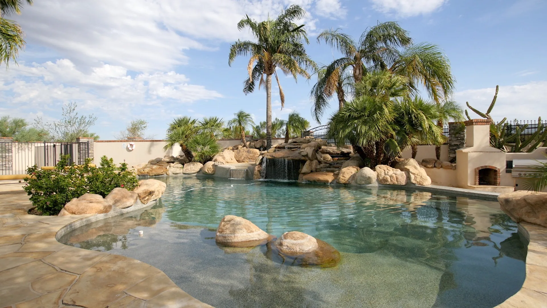 Free-form pool edged with boulders and palm trees under a blue sky, a stone fireplace behind