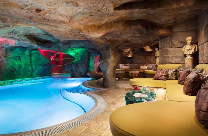 Indoor pool in a rock grotto with coloured lighting, yellow lounge seating and a classical bust