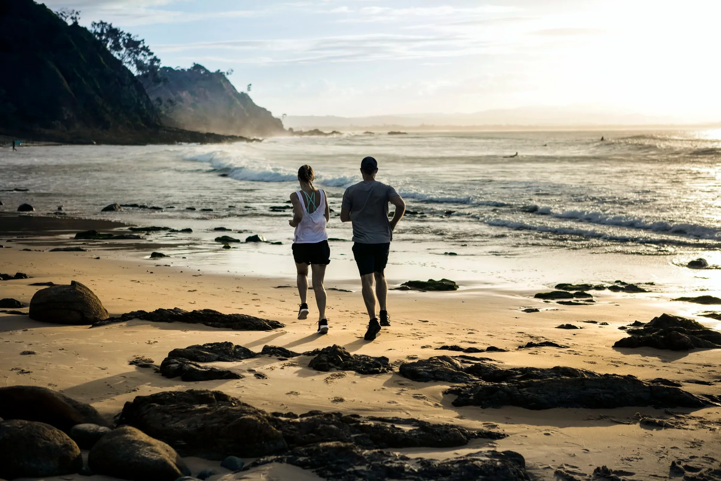 Two people walking along a rocky beach at sunrise with surf and a forested headland behind