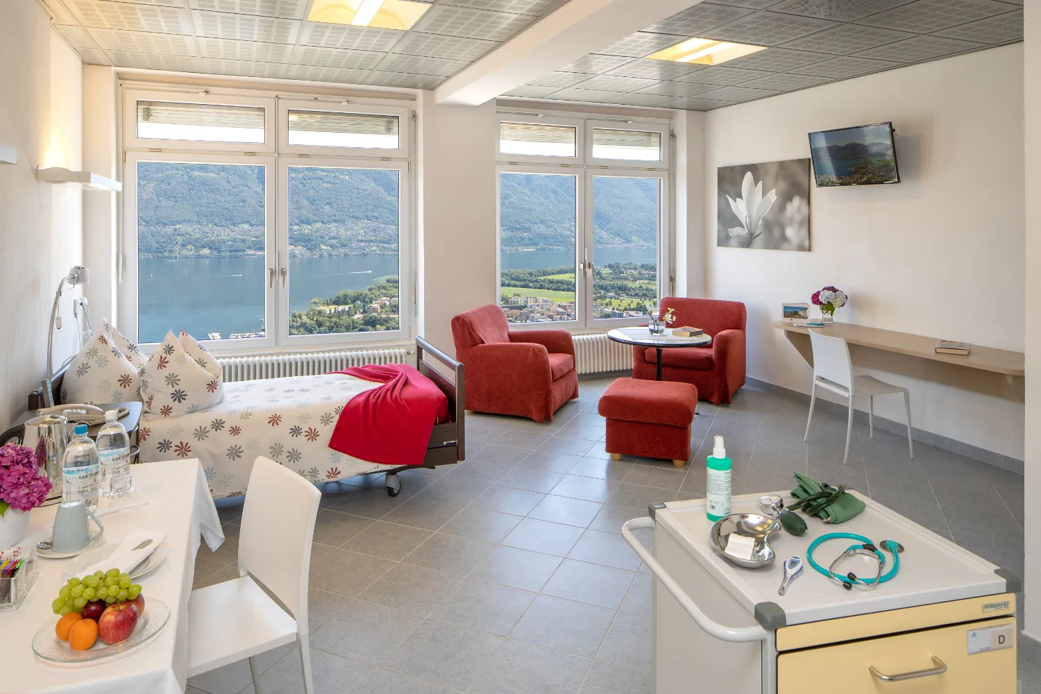 Third floor patient room with a bed, red armchairs, a desk and two windows facing the lake