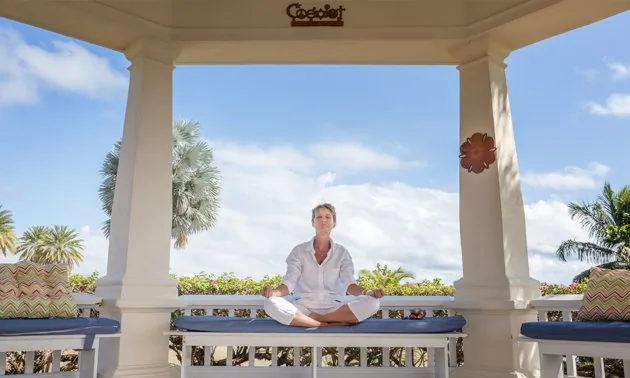 Person meditating on a cushioned bench inside a white gazebo with palm trees and blue sky
