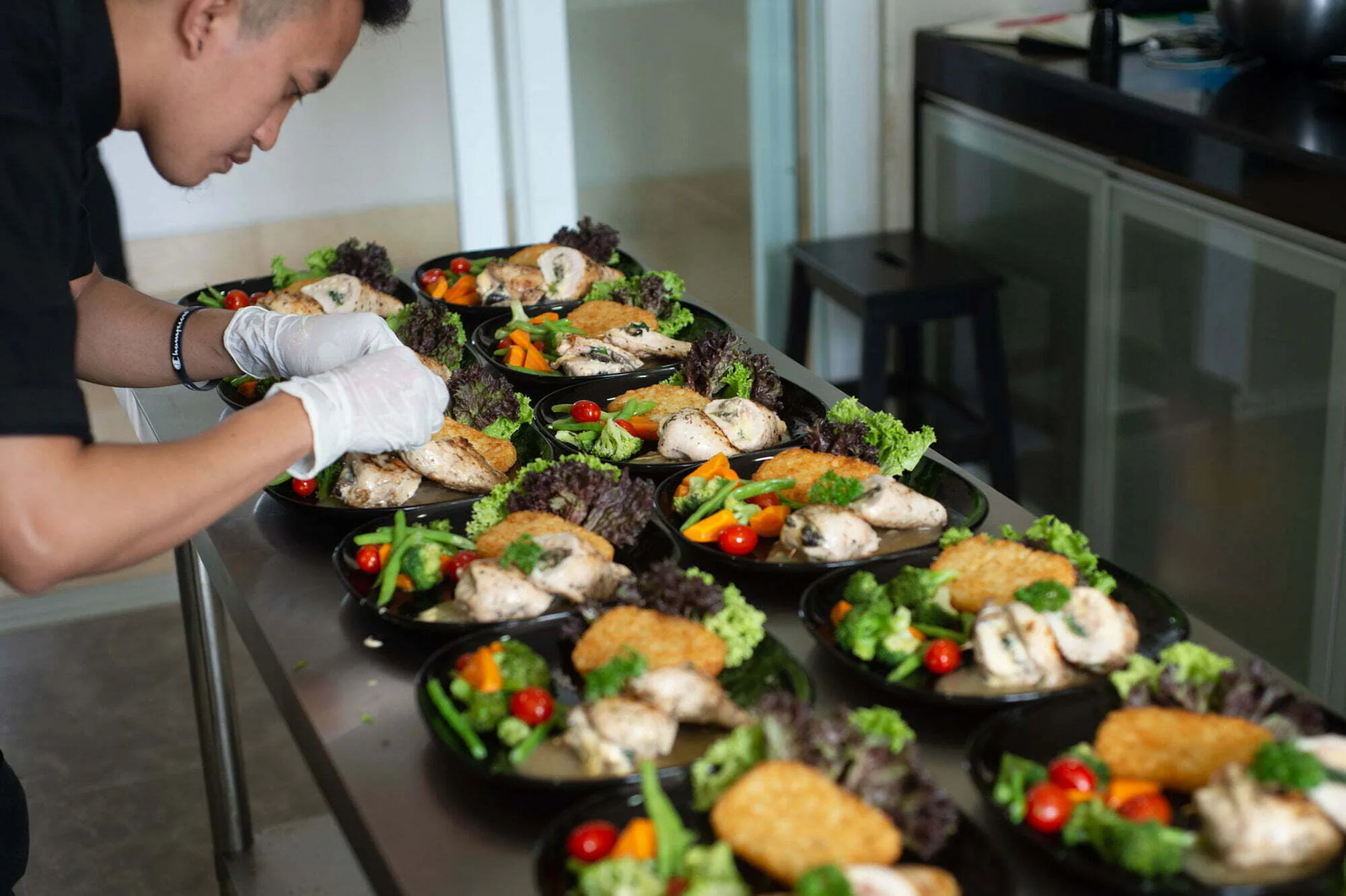 Gloved kitchen staff member plating rows of meals with salad, rice and grilled fish on a steel counter