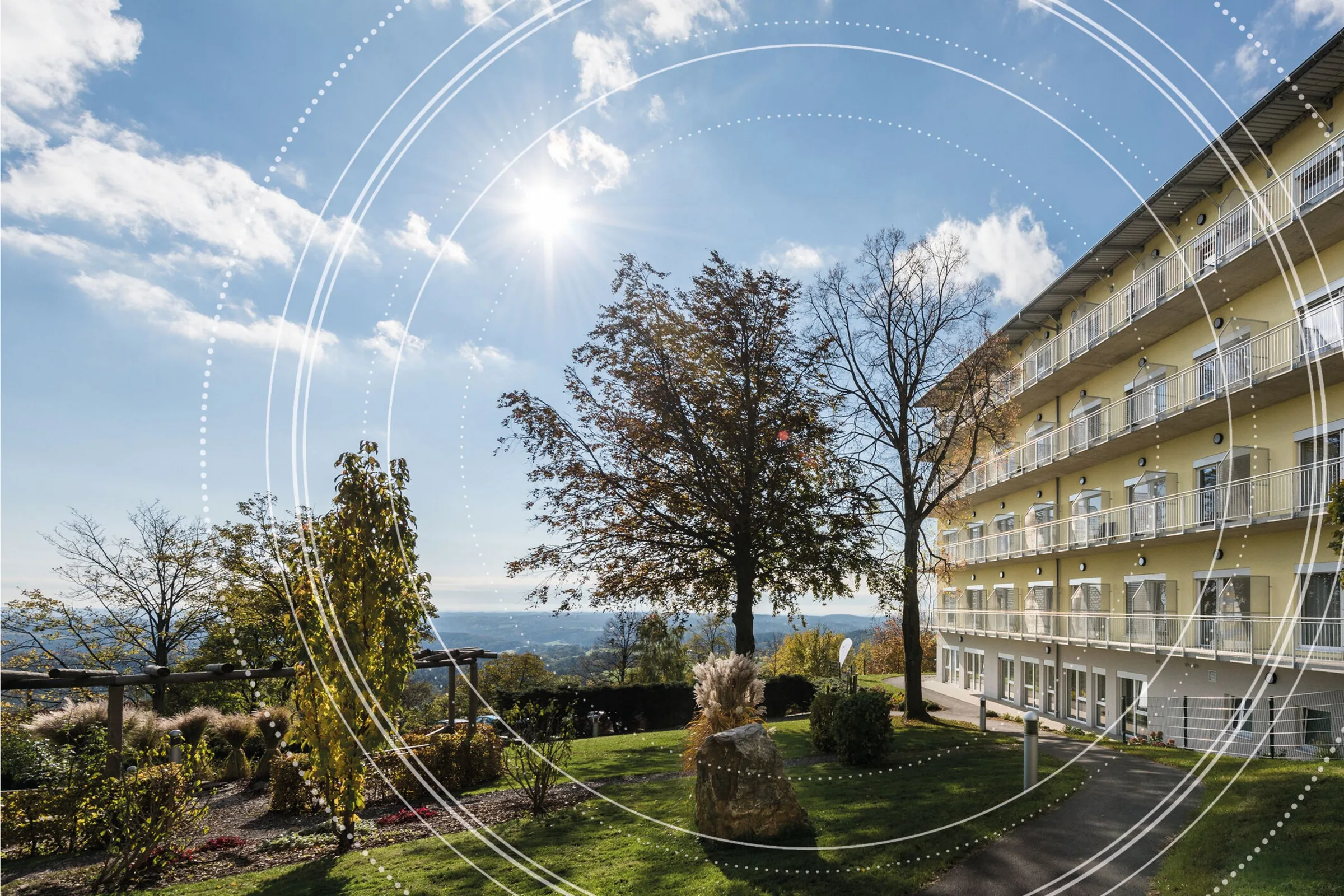 Multi-storey clinic building with balconies beside a sunny garden with trees and a wide hilly view