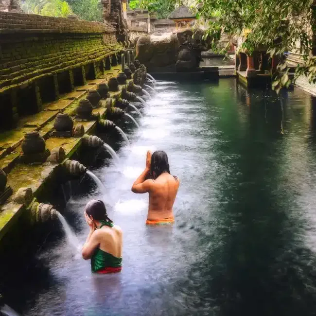 Two bathers in a stone temple pool fed by a row of carved spouts, surrounded by tropical trees