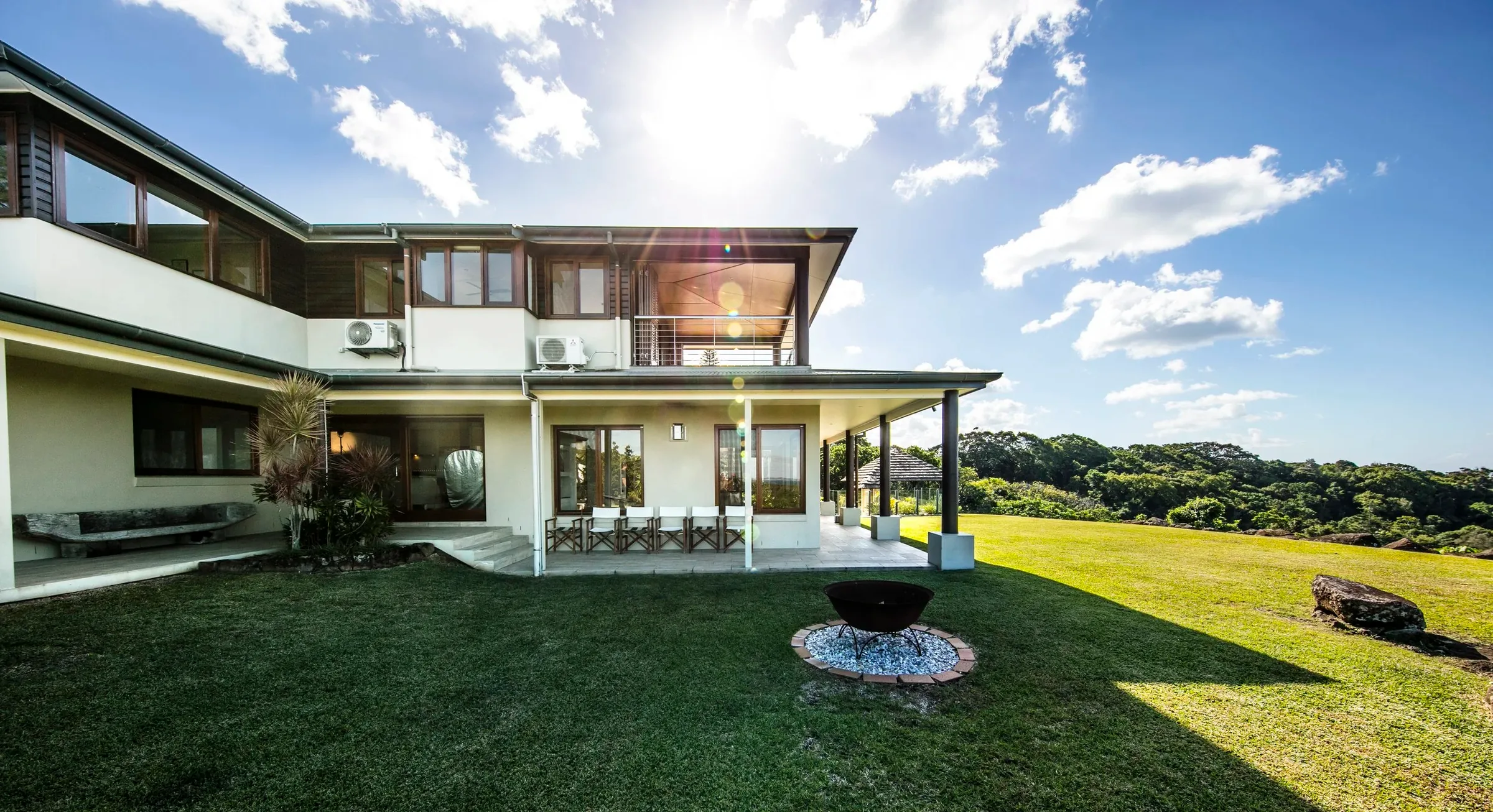 Two-storey white residence with wide verandas and balconies above a sunny lawn and fire pit