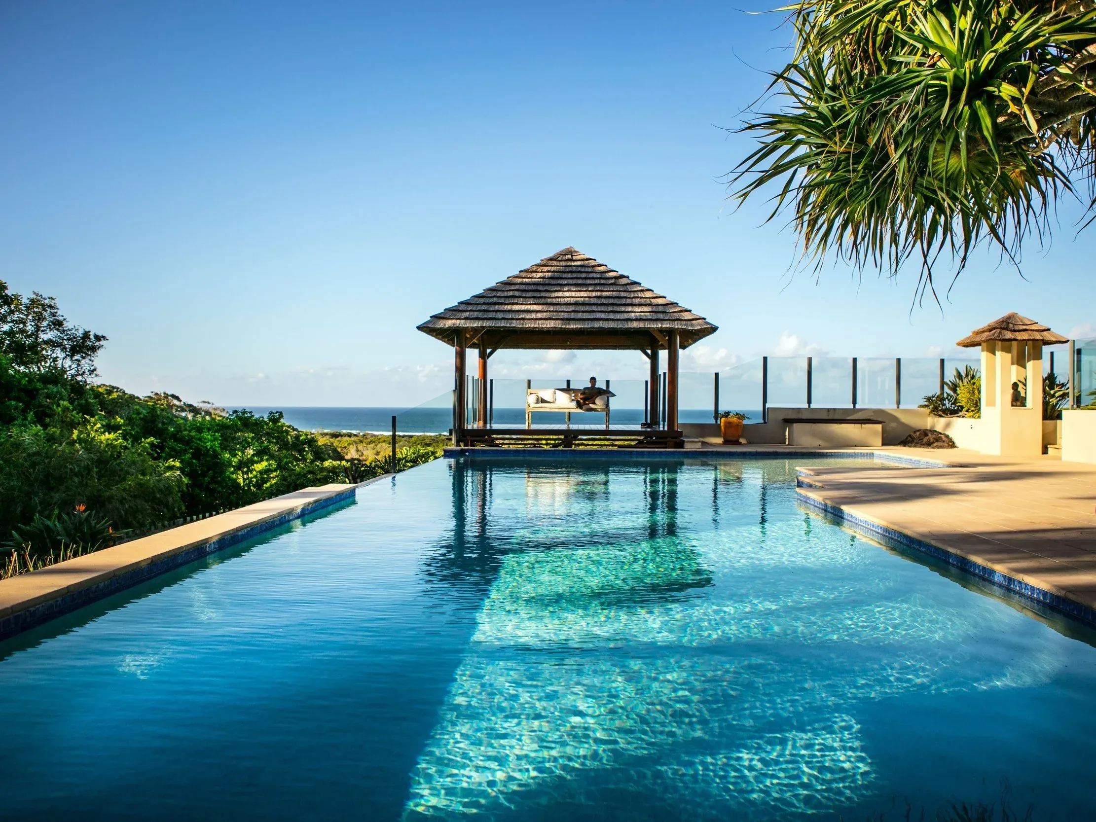 Infinity pool with a thatched gazebo at its far end, timber decking and the ocean on the horizon