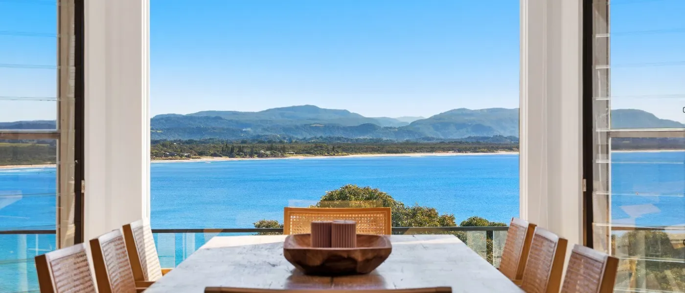 Dining table in front of open windows looking over a blue bay to hills on the far shore