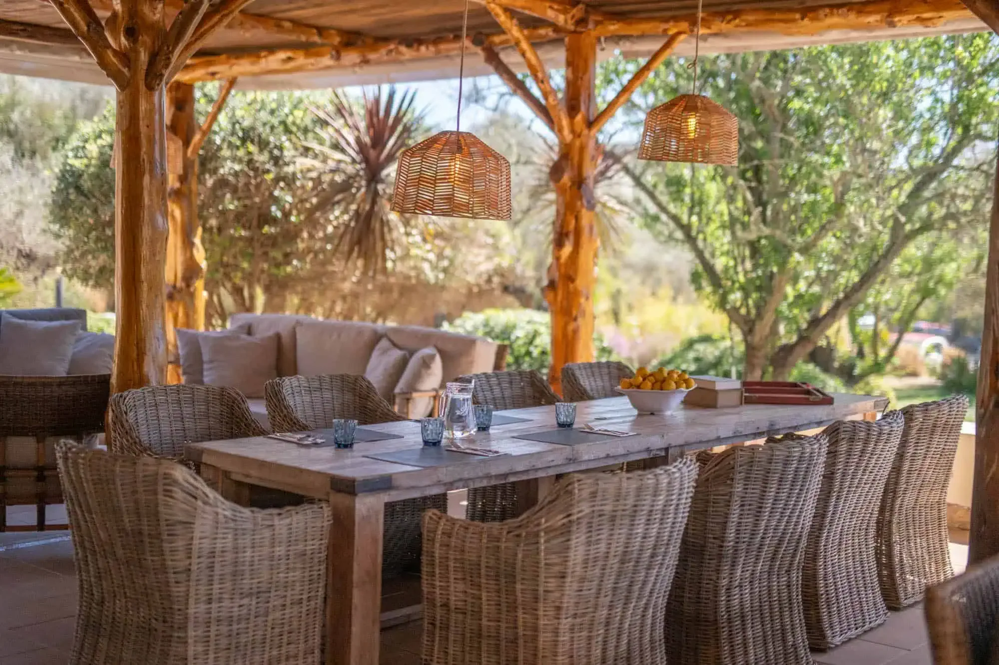 Long dining table with wicker chairs under a rustic timber pergola with hanging lamps and olive trees