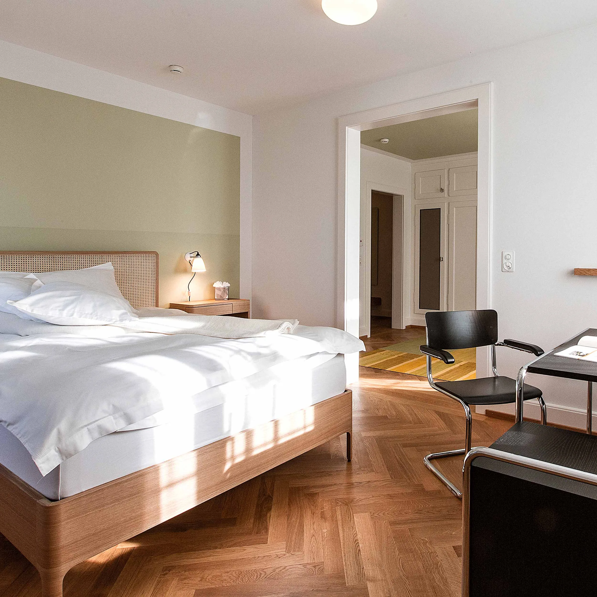 Bright client room with a wooden double bed, white linen, herringbone parquet and a desk chair