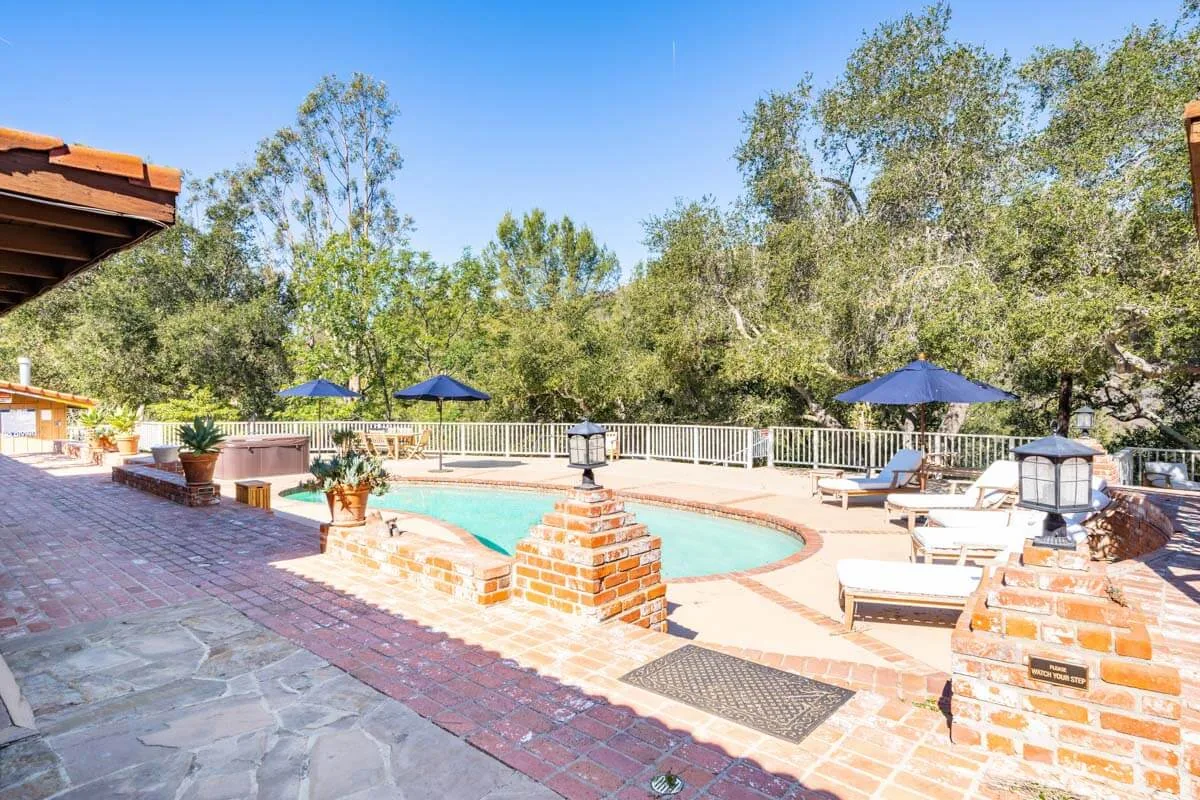 Brick pool terrace with blue umbrellas, sun loungers and a spa among oak trees
