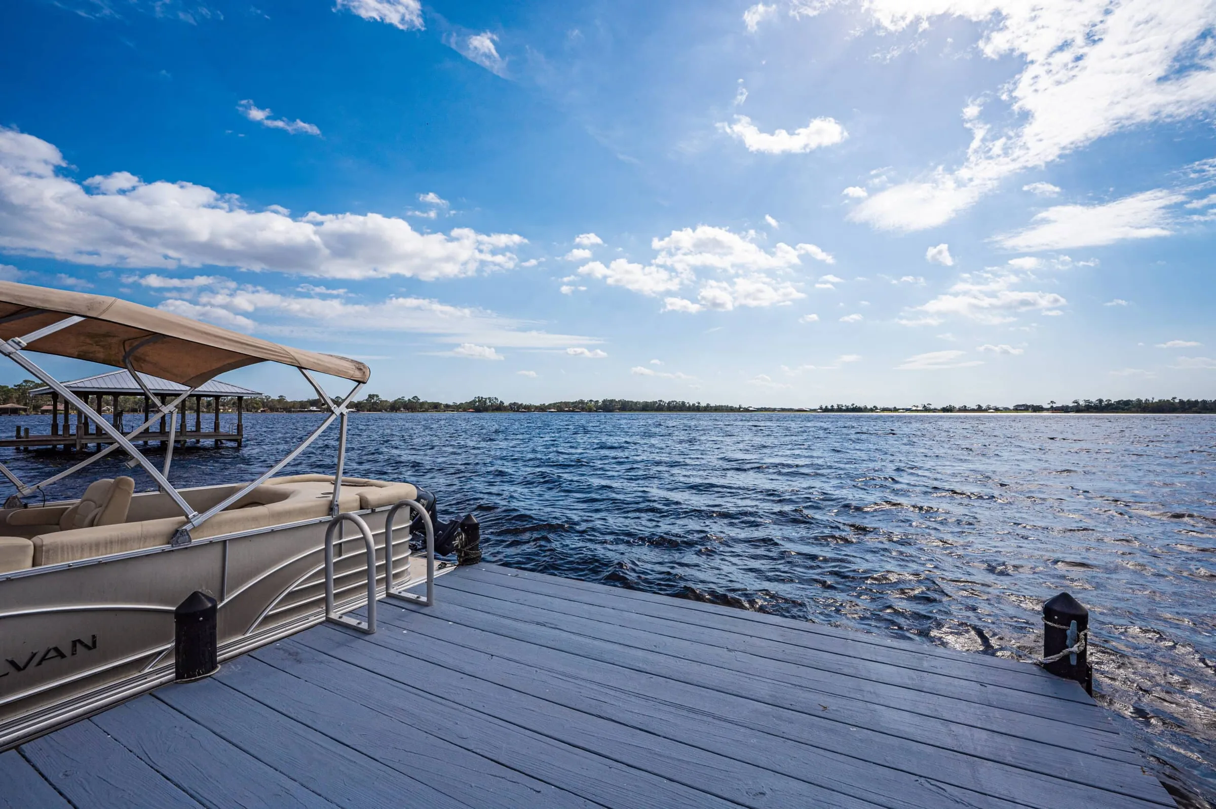 Wooden dock with a covered pontoon boat moored on a wide lake under a blue sky with clouds