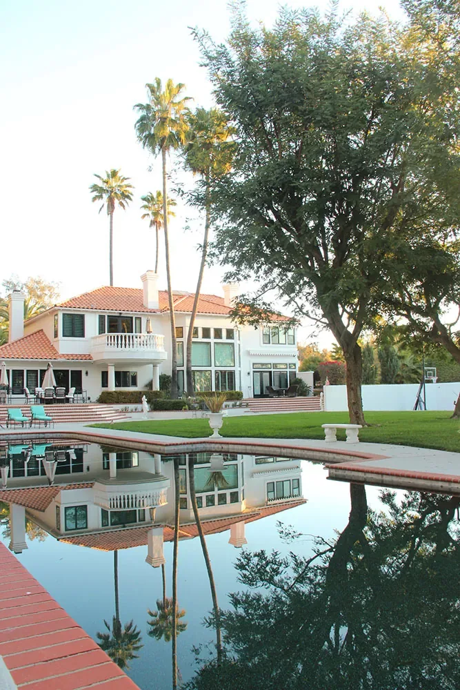 Still pool reflecting a white house with a red roof, tall palms and trees under a pale sky
