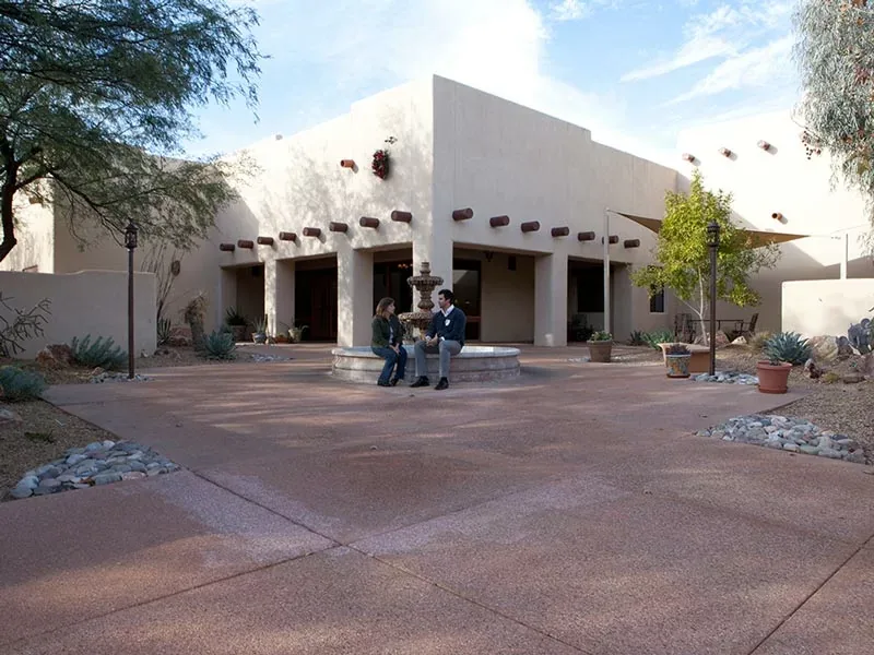 White adobe style main building with a covered walkway, fountain and paved courtyard under blue sky