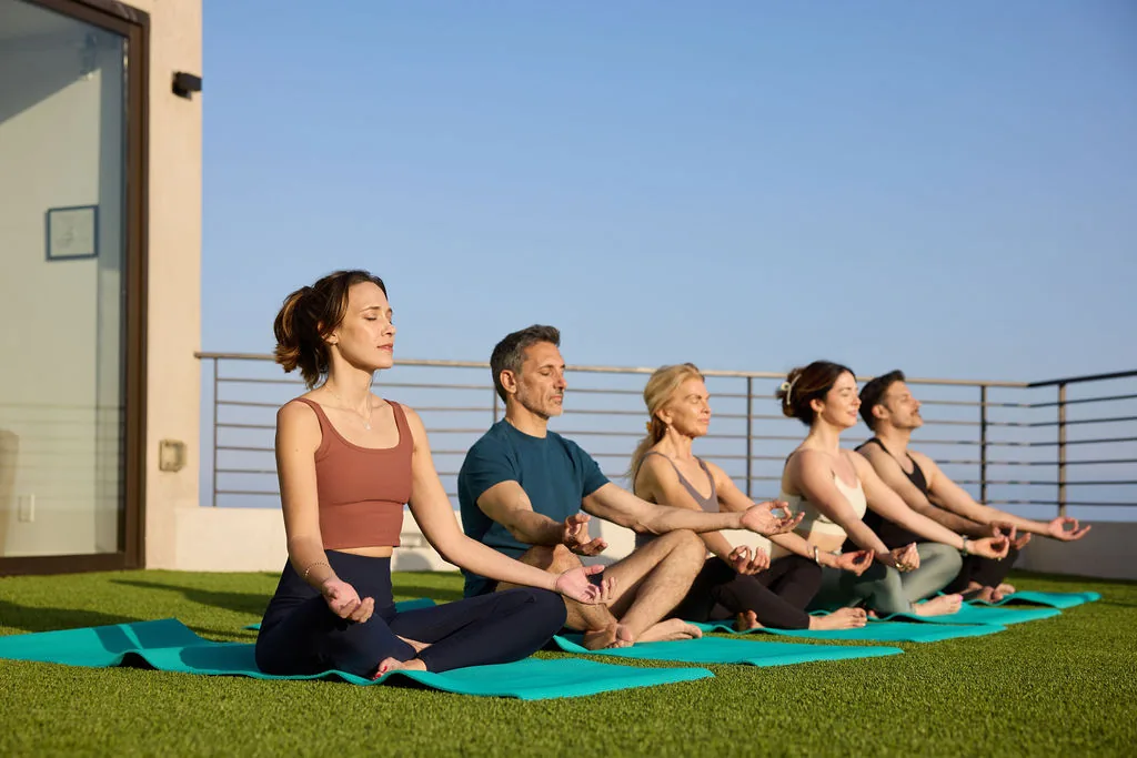 Five people meditating on yoga mats on an artificial lawn terrace with a glass railing and blue sky