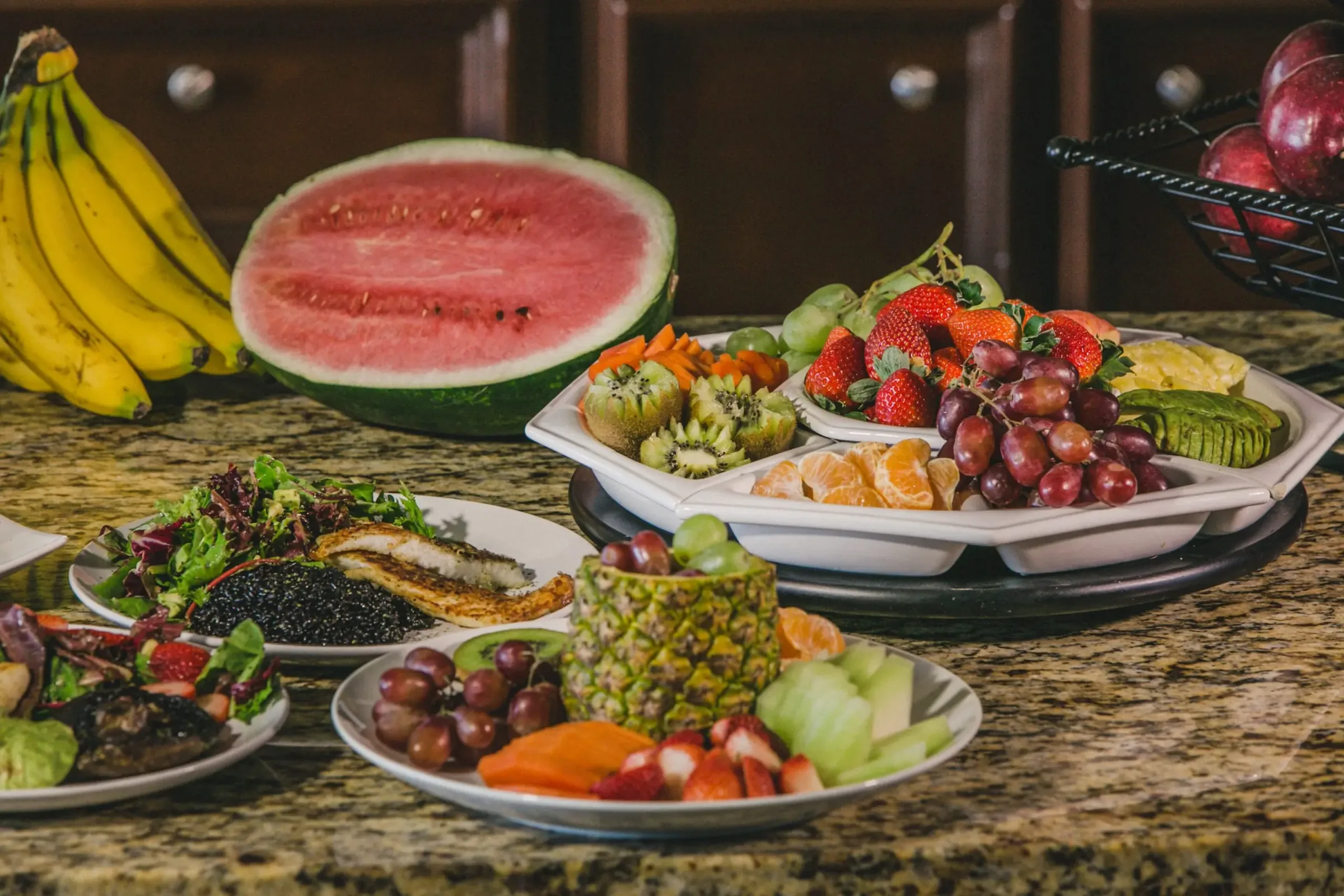 Kitchen counter laid with platters of fresh fruit, watermelon, salad and grilled fish