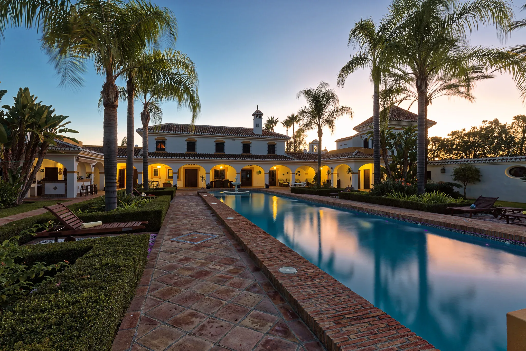 Narrow pool reflecting the lit main building and tall palms at dusk, with a paved path alongside