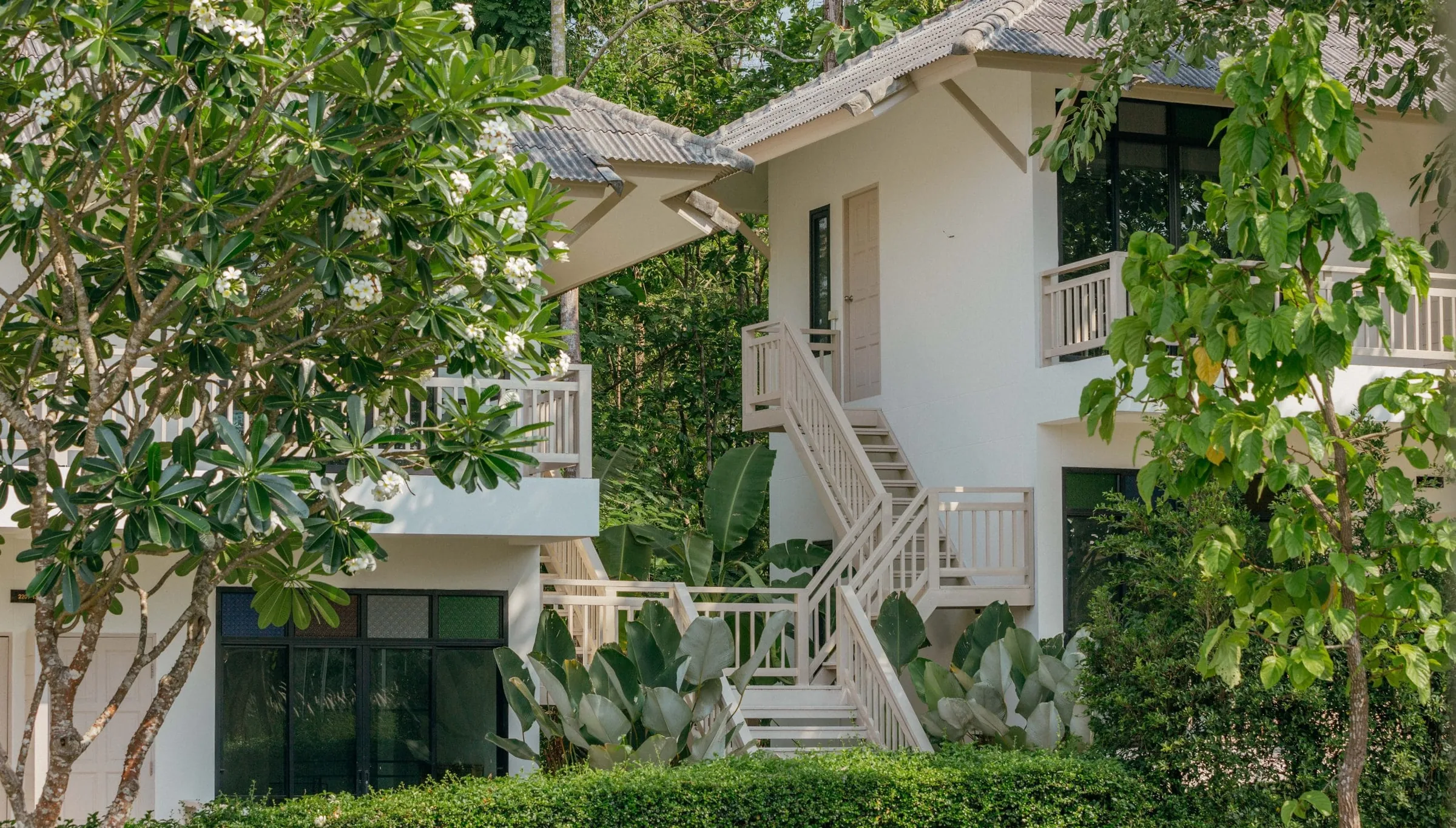 White two-storey residential buildings with balconies and outdoor stairs, framed by leafy trees