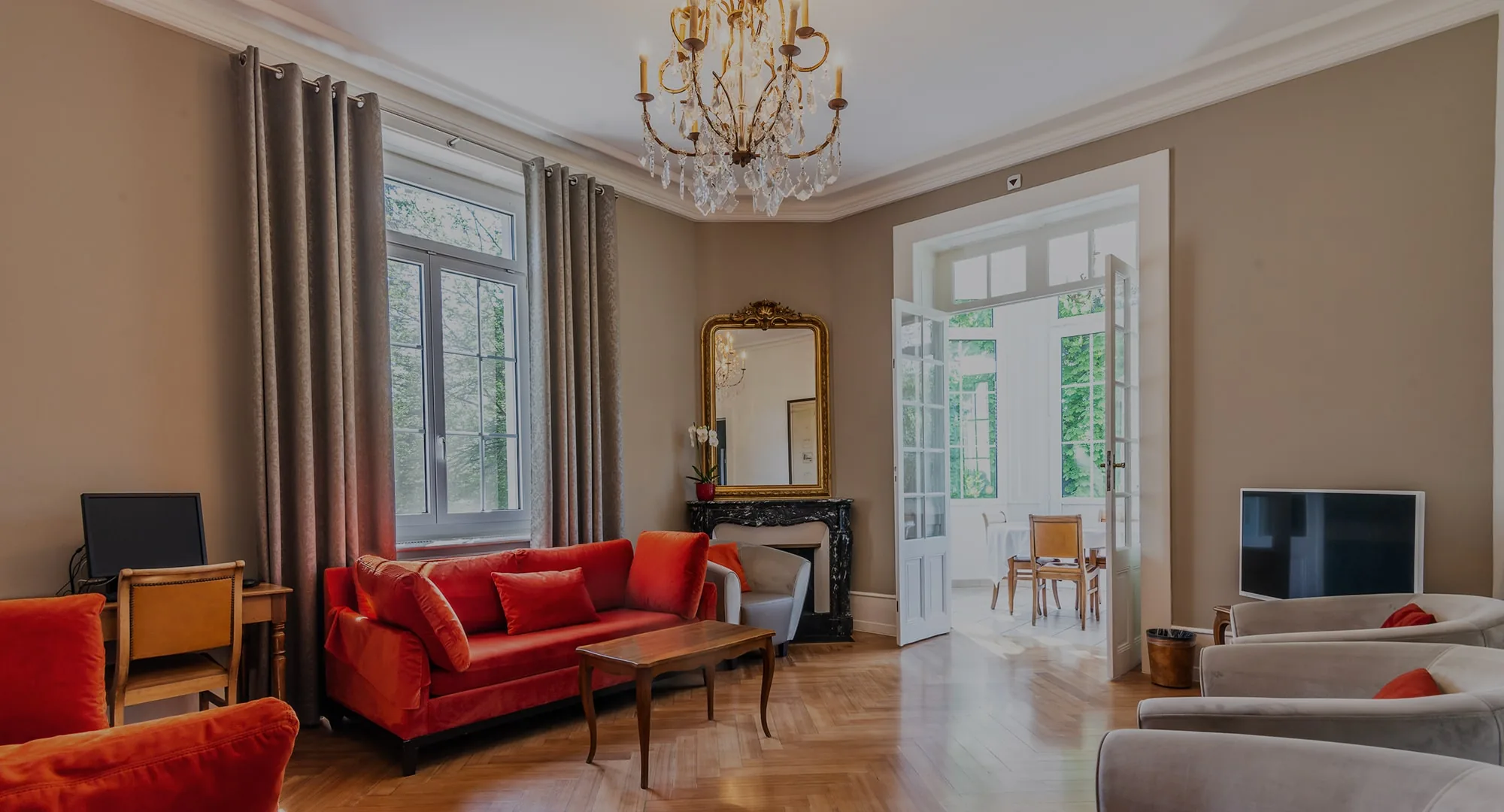 High-ceilinged salon with a crystal chandelier, red sofas, a gilt mirror and tall windows onto the garden