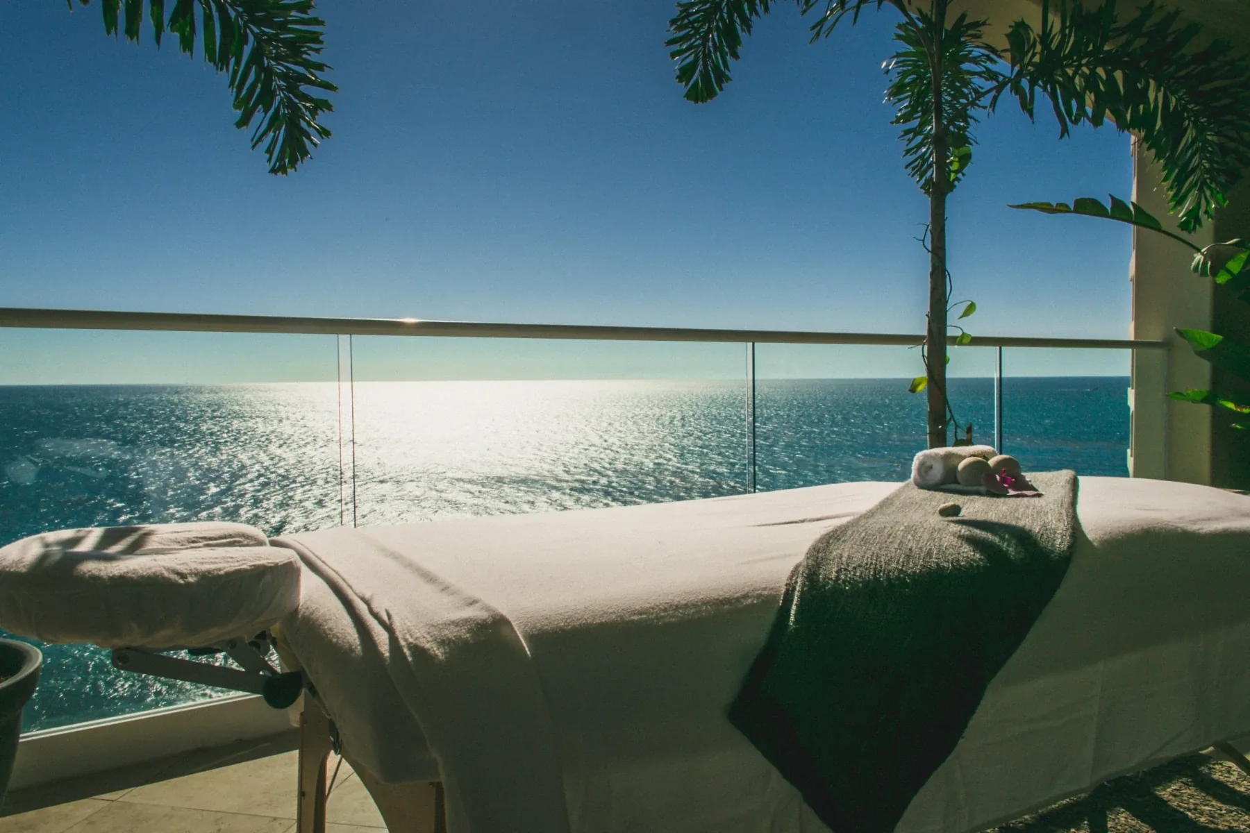 Massage table with folded towels on a glass-railed terrace overlooking the sunlit ocean