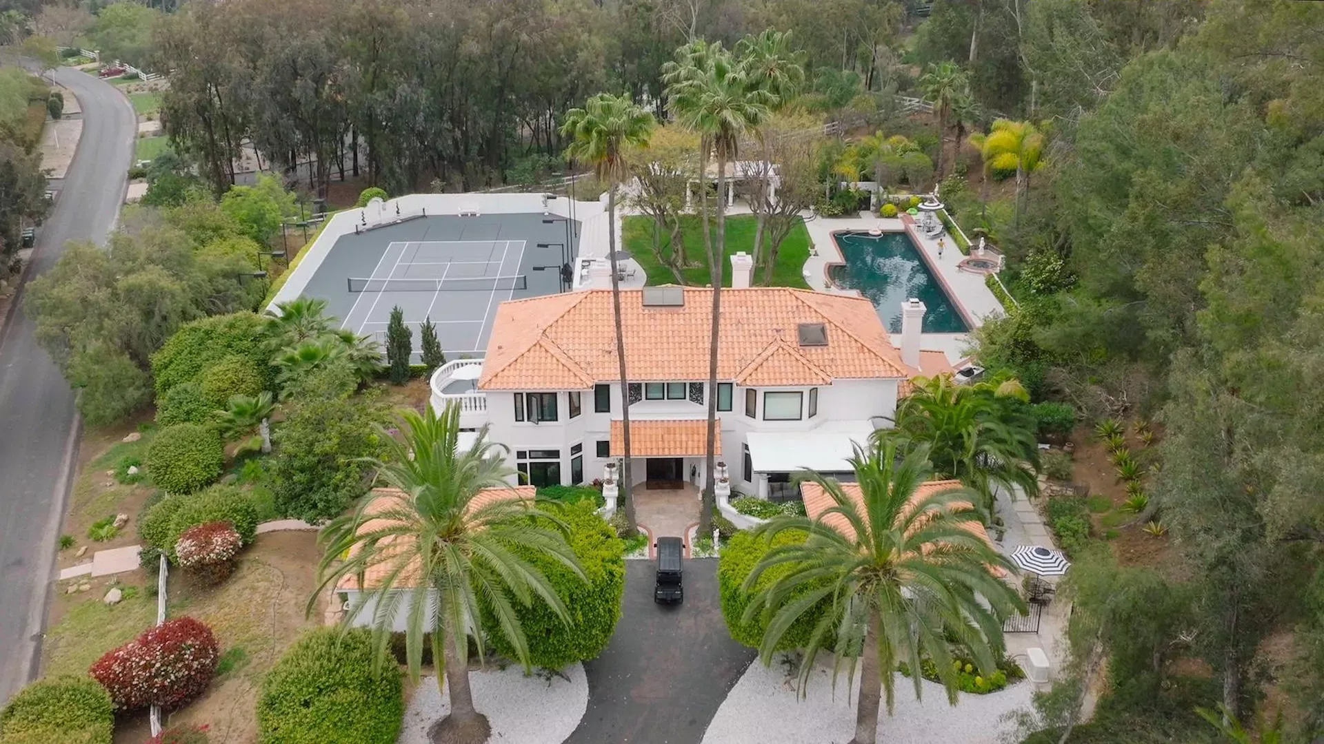 Aerial view of a white villa with a tennis court, pool and palm-lined driveway among trees in Encinitas