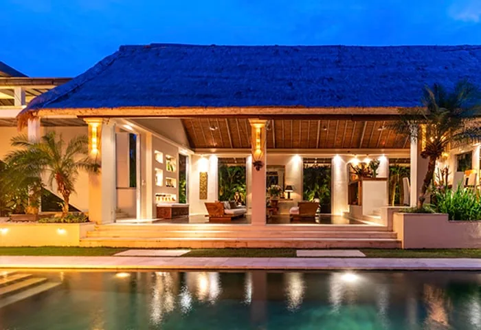 Lit open-sided villa pavilion with a dark pitched roof above a pool at dusk in Bali
