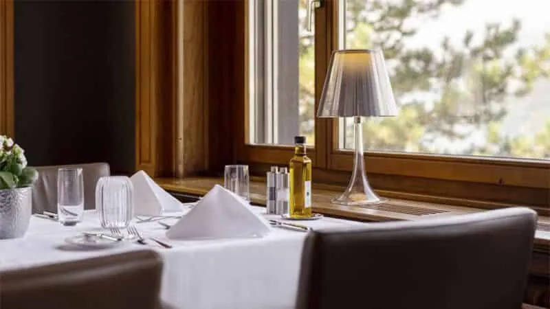 Restaurant table laid with white linen and glasses beside a large window with a table lamp and trees outside