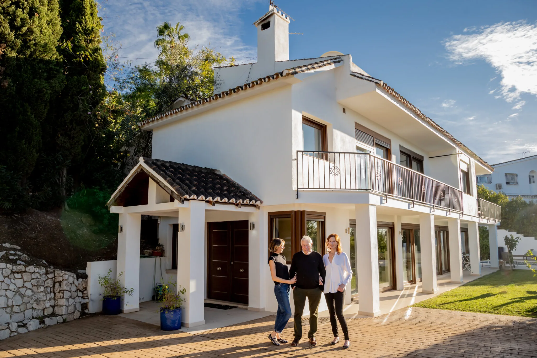 White two-storey Andalusian villa with a tiled porch and balcony, three people standing at the front door