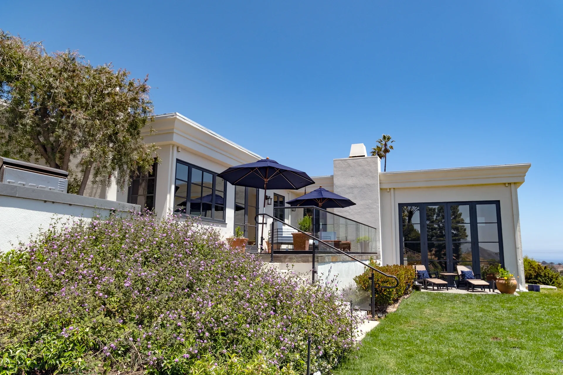 White single storey house with patio umbrellas above a flowering hillside garden and lawn in Malibu