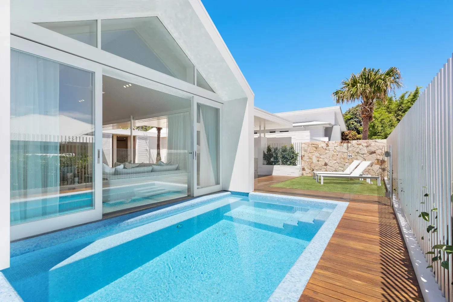 Blue lap pool beside a white gabled residence with timber decking and a stone wall