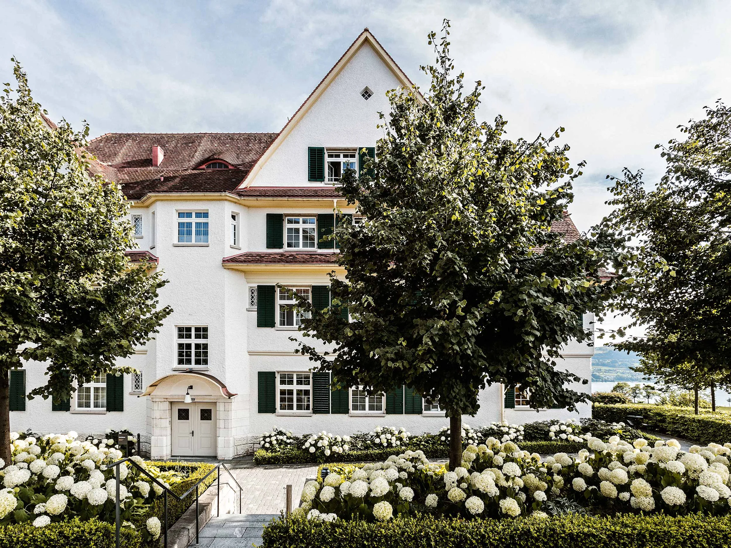 White gabled main clinic building behind trees and beds of white hydrangeas under a pale sky