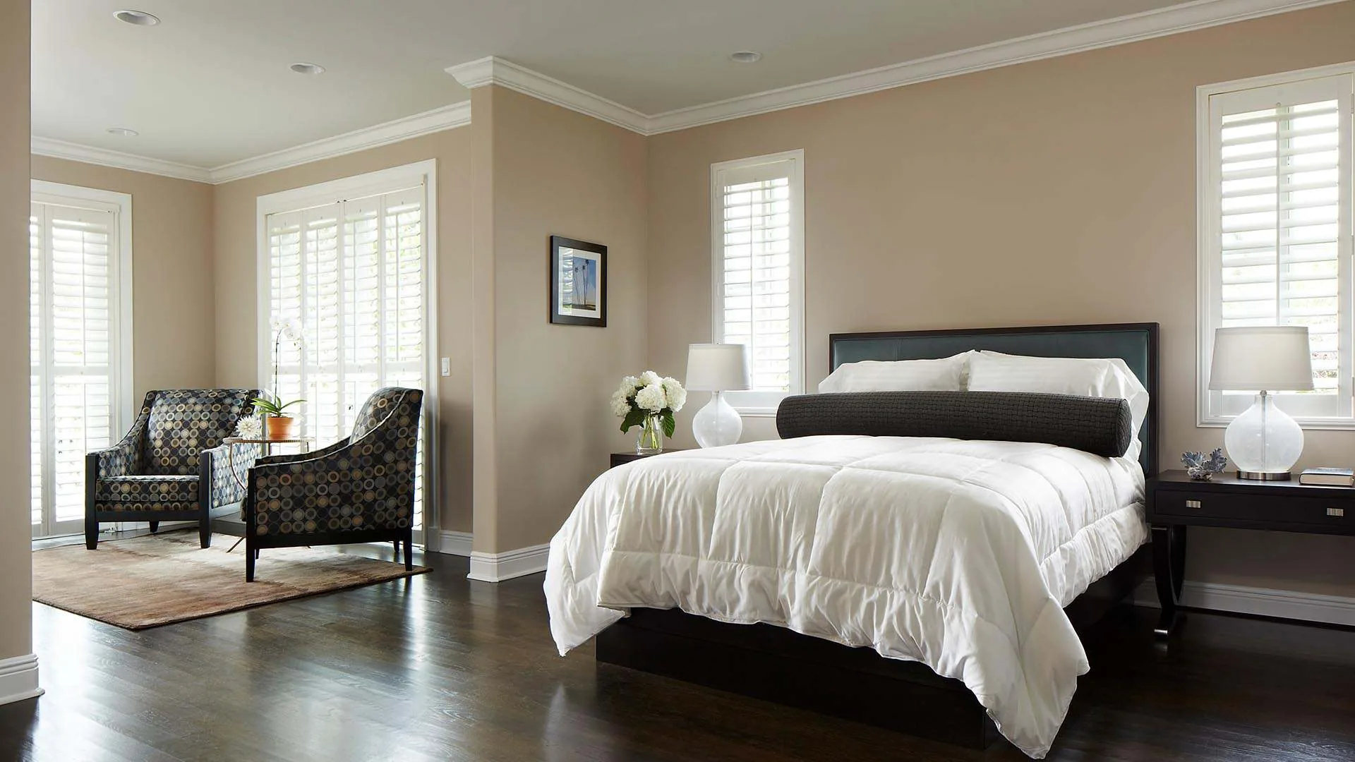 Bedroom with white bedding, dark wood floor, shuttered windows and two patterned armchairs