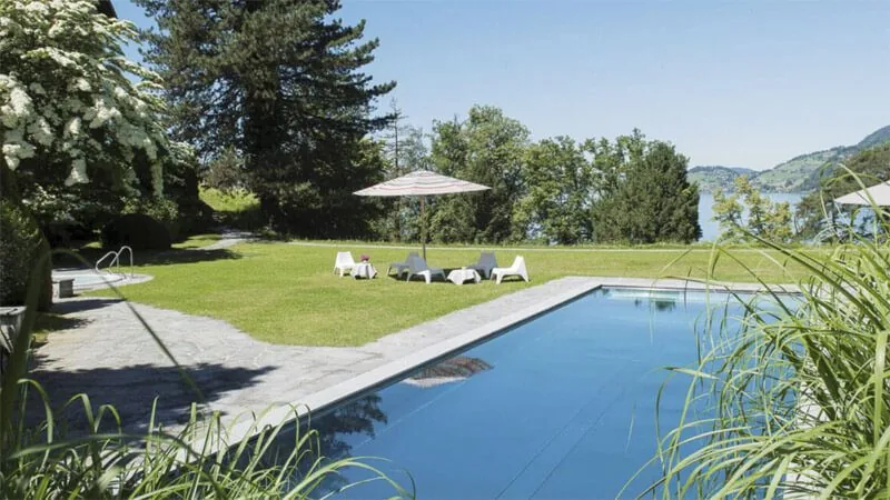 Outdoor swimming pool beside a lawn with parasols and sun loungers, framed by tall trees and lake hills
