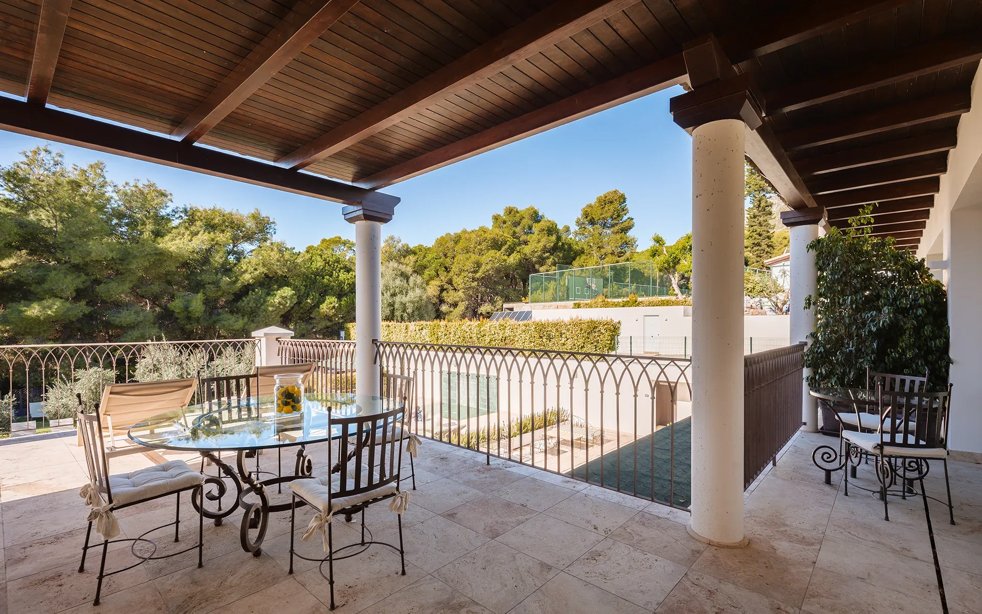 Covered terrace with a wooden beam ceiling, a round table and chairs and pine trees beyond