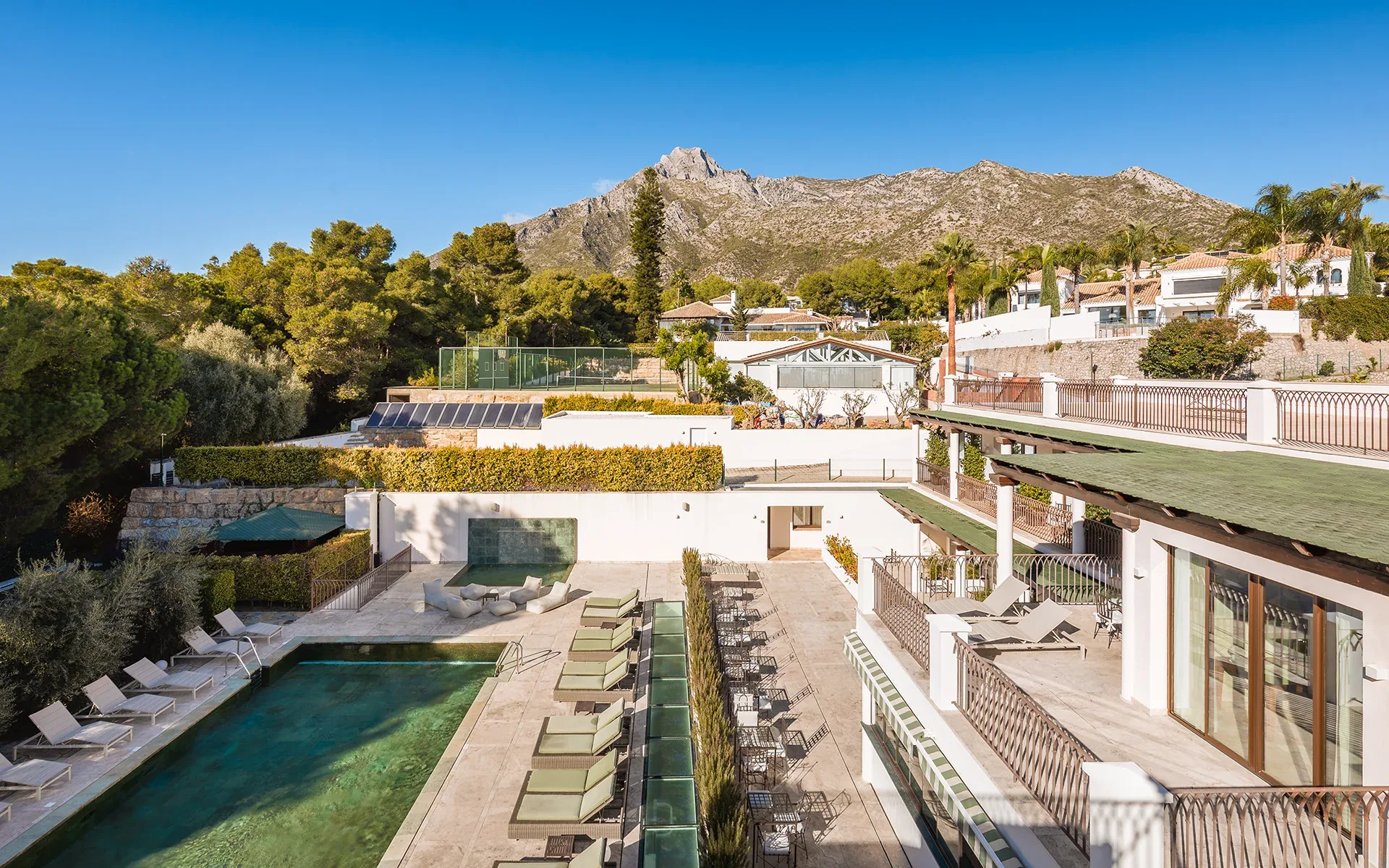 Elevated view of a pool, sun loungers and gardens with a rocky mountain under blue sky