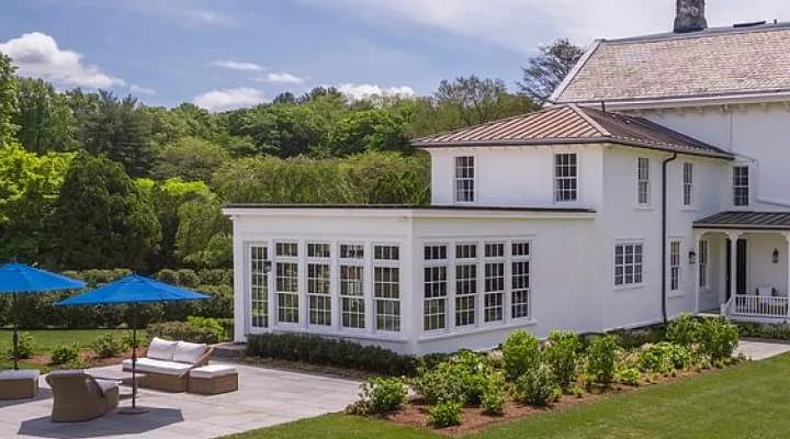 White clapboard house with a glass sunroom, patio umbrellas and clipped hedges on a green lawn